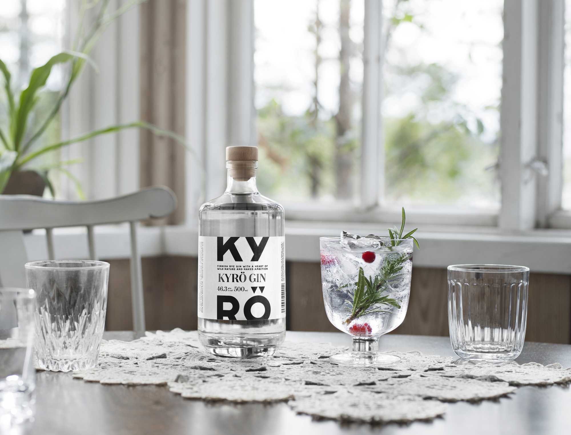 A bottle of Kyrö Gin standing on a dining table in a light-filled room next to an Iittala cocktail glass filled with Gin & Tonic, rosemary, and cranberries. Horizontal. 