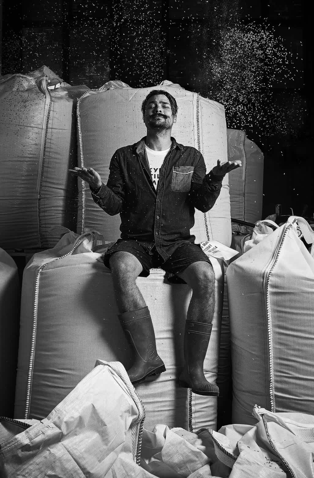 Black and white image: head distiller Kalle Valkonen sit atop bags of rye, tossing kernels into the air. 