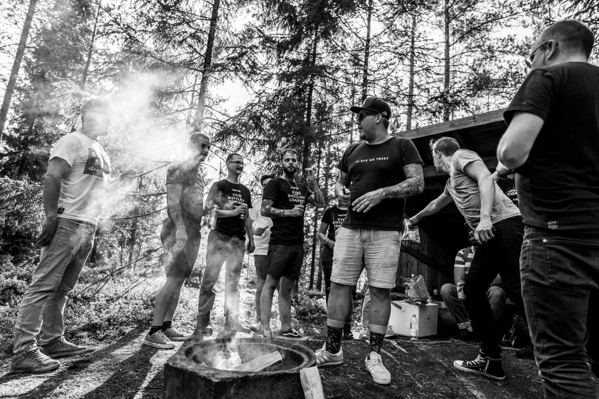 Group of people standing in the middle of a Finnish forest in a very bright sunlight. Next to them there is a campfire.
