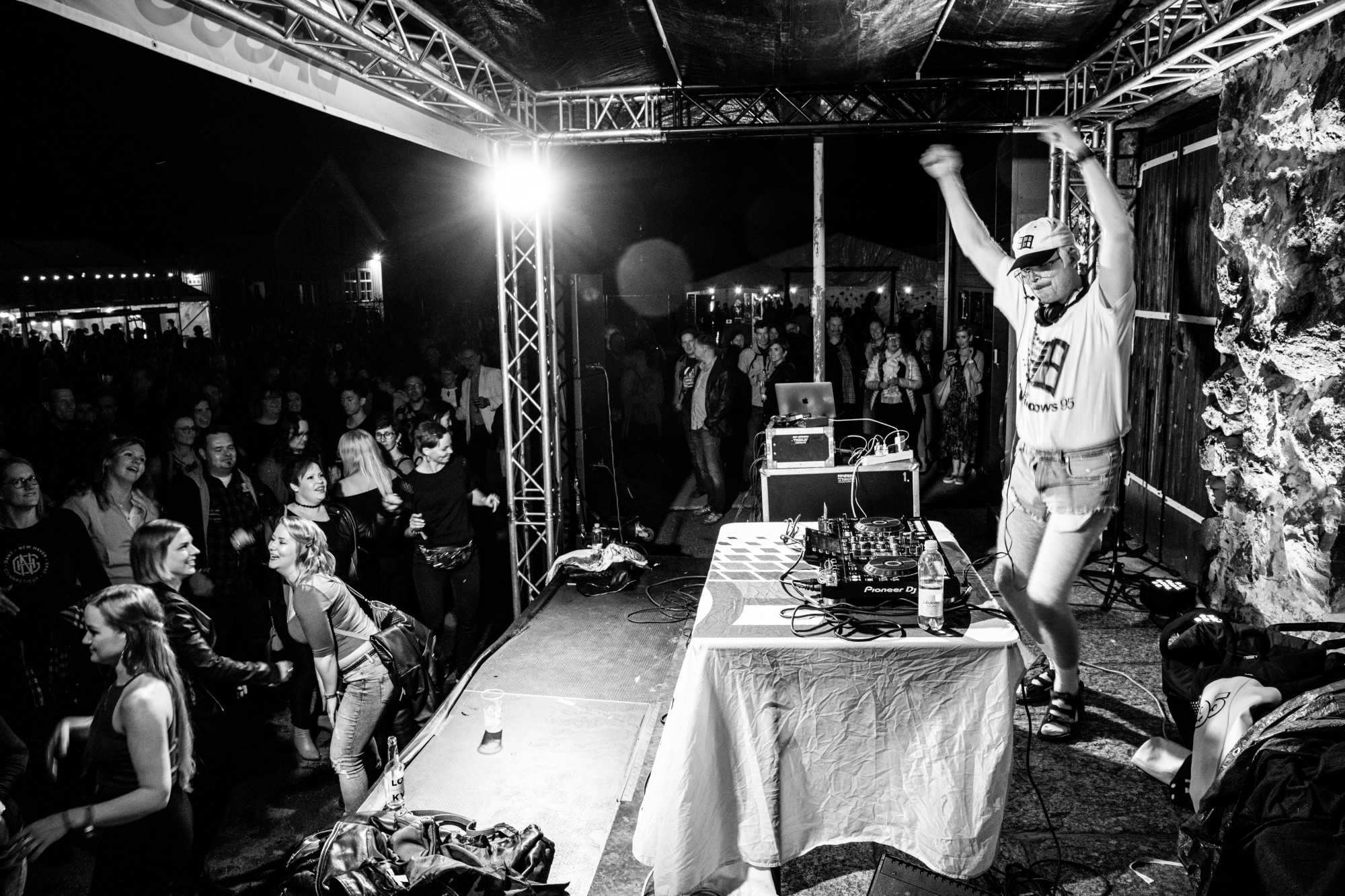 A black and white image of a DJ standing in front of a crowd on a podest. Everyone is dancing. 