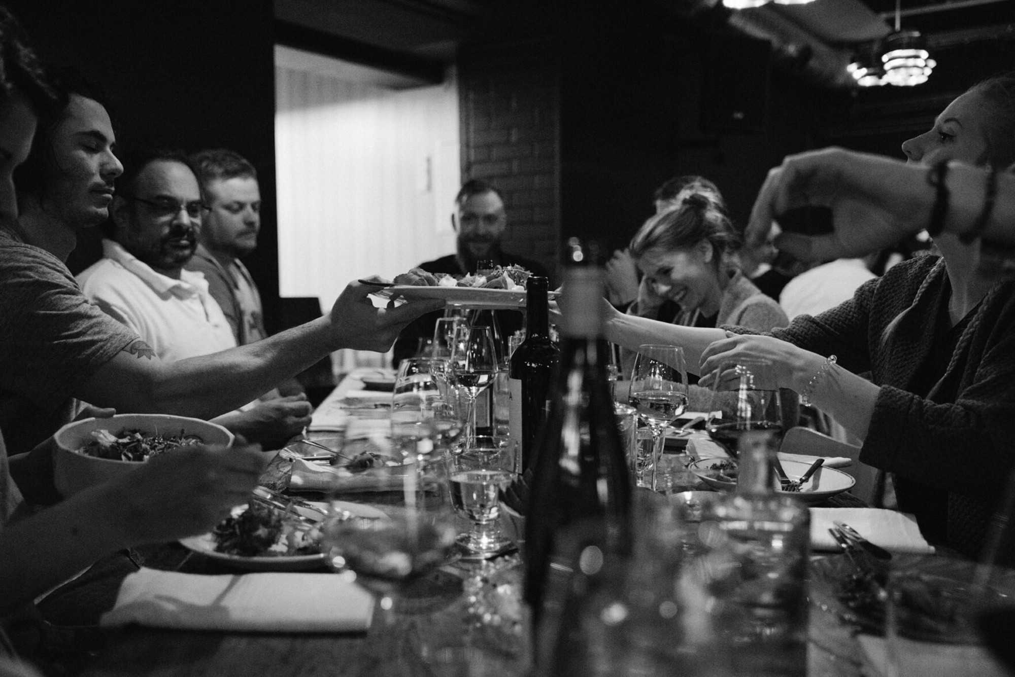 Black and white image of a group of people sat across each other on a long, communal table sharing plates and enjoying good drinks. 