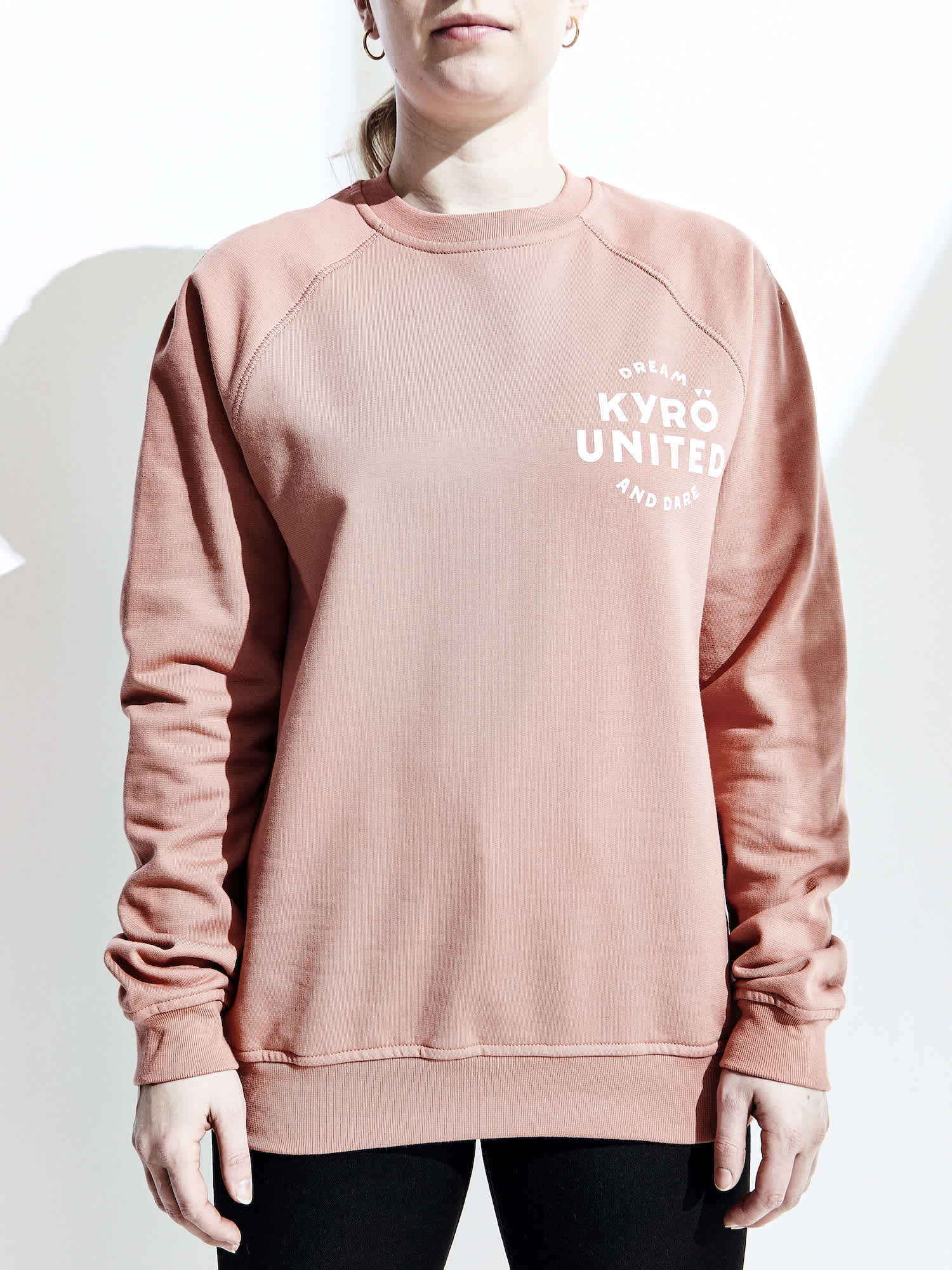 A white, female model wearing a salmon-colored pink sweater made in collaboration with Kyrö and R-Collection. Logo on upper left chest. 