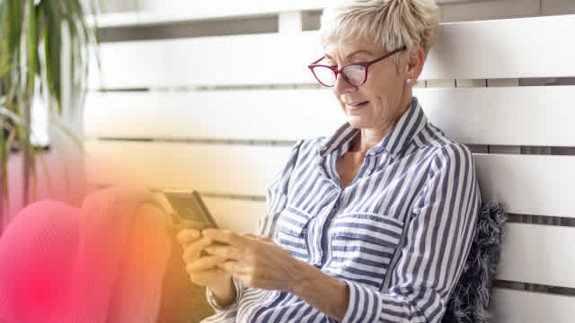 Smiling older woman in striped shirt and red glasses using smartphone to play online casino games, representing mobile gambling and gaming analytics in a relaxed home setting.
