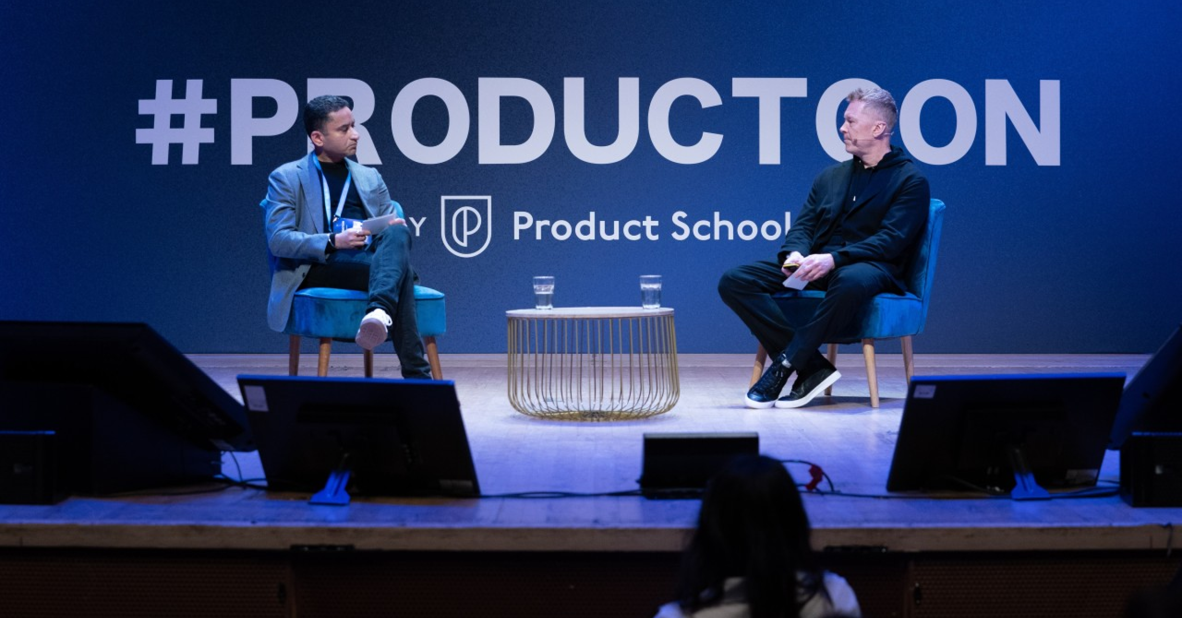 Two speakers on stage in blue chairs at Product School event, with #PRODUCTCON backdrop and small table between them.