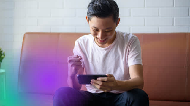 Mobile gamer enjoying gameplay on a handheld device, seated casually against a white brick wall with vibrant purple and pink gradient lighting effects adding a modern gaming atmosphere.