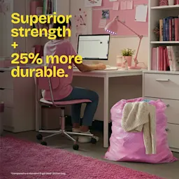A room decorated with pink accents, a young woman sitting at a desk with a pink trash bag next to her. The words "Superior strength + 25% more durable." in yellow
