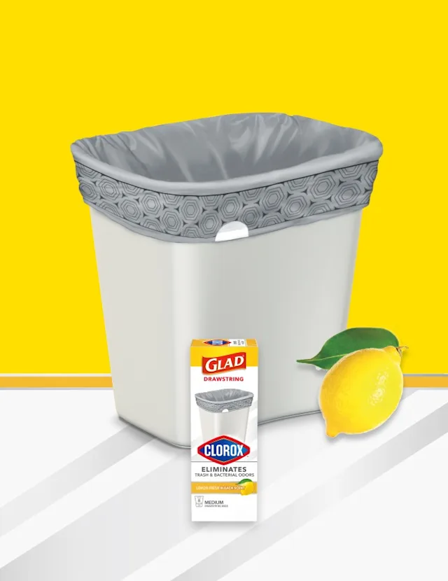 White plastic trash can with gray trash bag inside, yellow and white background with lemon and box of Glad bags in front.