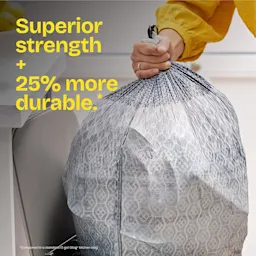 A person pulling a gray-color trash bag out of a trash can. The words "Superior strength + 25% more durable." in yellow