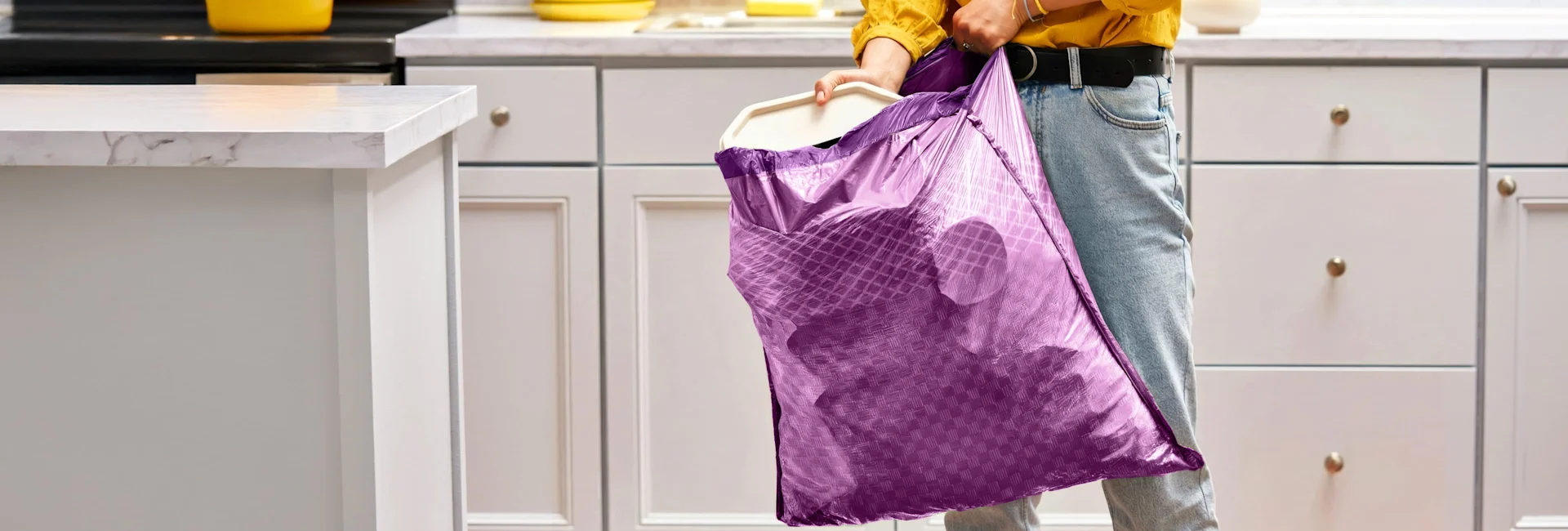 A person in jeans and a yellow blouse pulling a purple trash bag out of a trash can