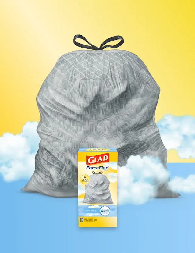 Gray trashbag with black handles on a background of yellow and blue with clouds. A box of Glad trash bags sits in front.