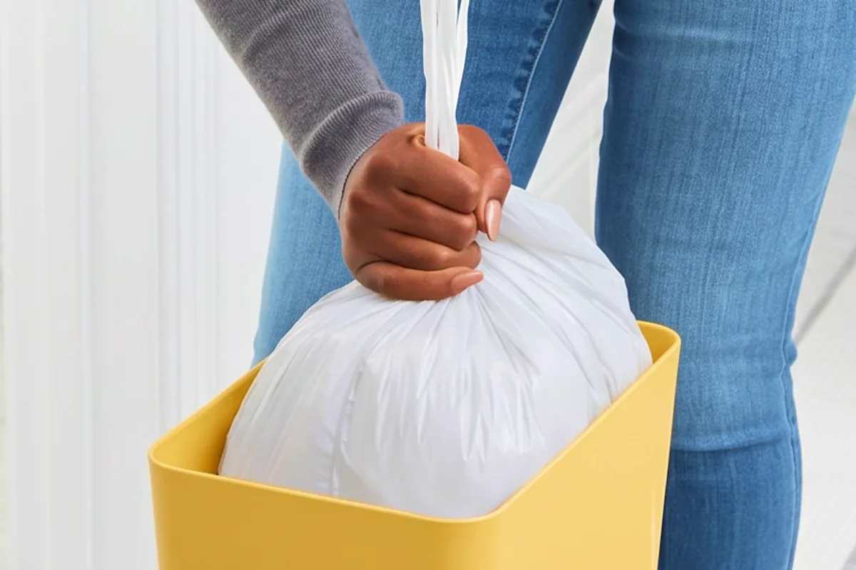 Hand lifting a tied white trash bag from a wastebasket for easy disposal