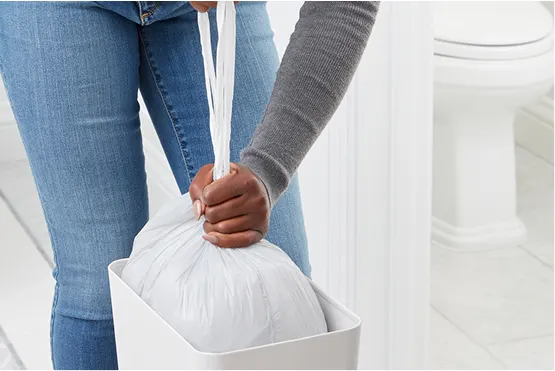 Person in gray shirt and blue jeans in a bathroom tying a knot in a small trash bag