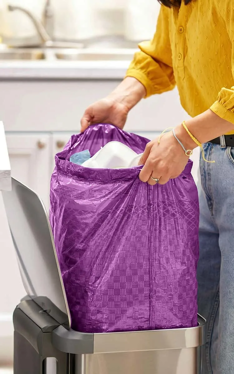 A person in jeans and a yellow blouse pulling a purple trash bag out of a trash can