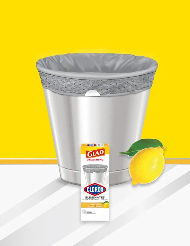 Small metal waste basket with gray trash bag inside on a yellow and white background. A lemon is in the lower right corner and a box of Glad small trash bags is in the front.