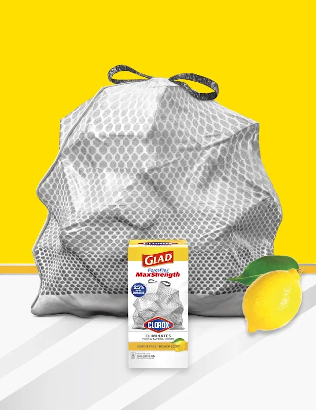 Gray trash bag with Glad package in front, yellow and white background with a lemon in the lower right corner.