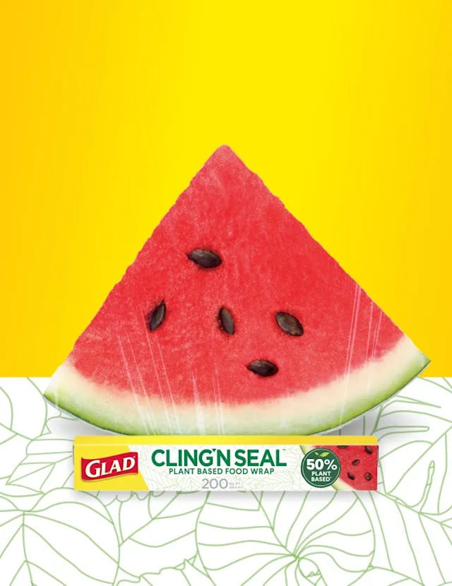 A quarter-slice of watermelon wrapped with Cling'N Seal on a yellow background with white tablecloth with green leaf drawings. A box of Glad Plant-Based Cling'N Seal sits in front.