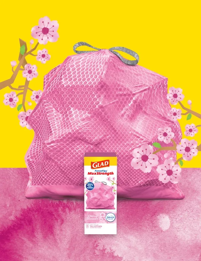 Pink trashbag with gray handles on a background of yellow and pink with cherry blossom illustrations. A box of Glad trash bags sits in front.