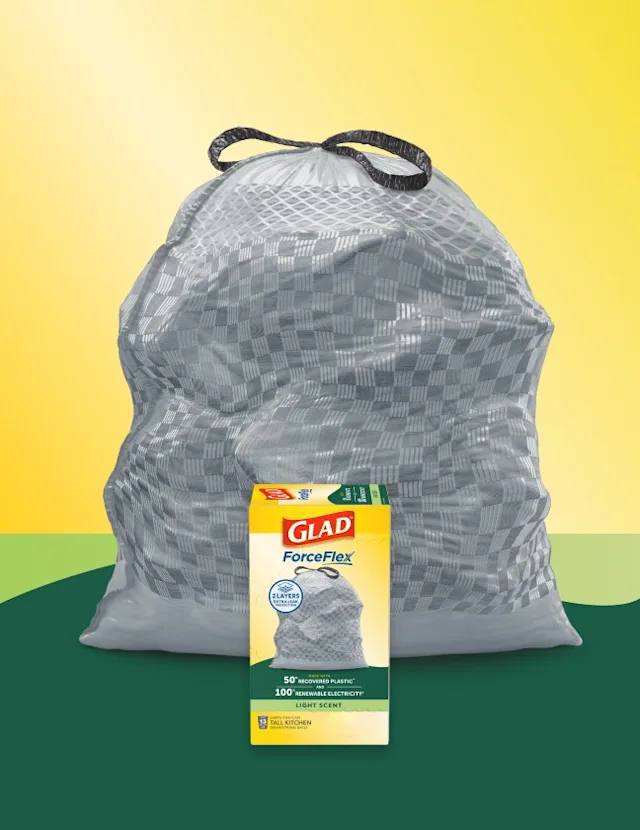 Gray trashbag with black handles on a background of yellow and green. A box of Glad trash bags sits in front.