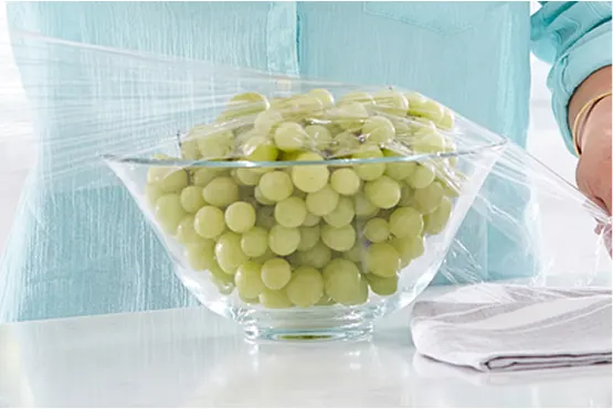 Person in light blue shirt putting plastic wrap over a glass bowl of green grapes