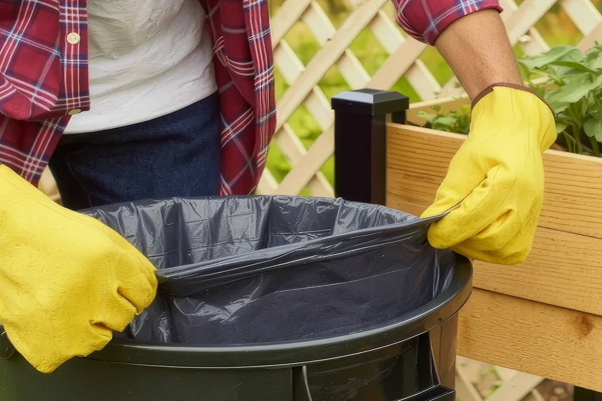 Person securing a black trash bag around an outdoor trash can with gloved hands
