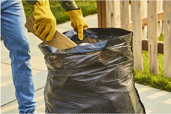 Person outside in yard, in blue jeans and yellow work gloves putting wood and debris in a black trash bag.