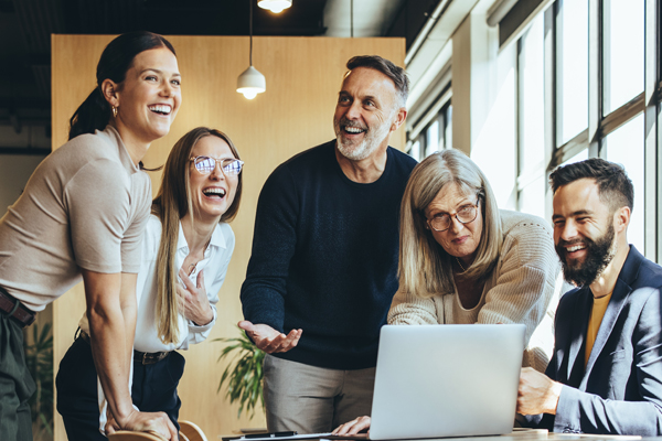 Five employees standing and smiling around a laptop THUMBNAIL