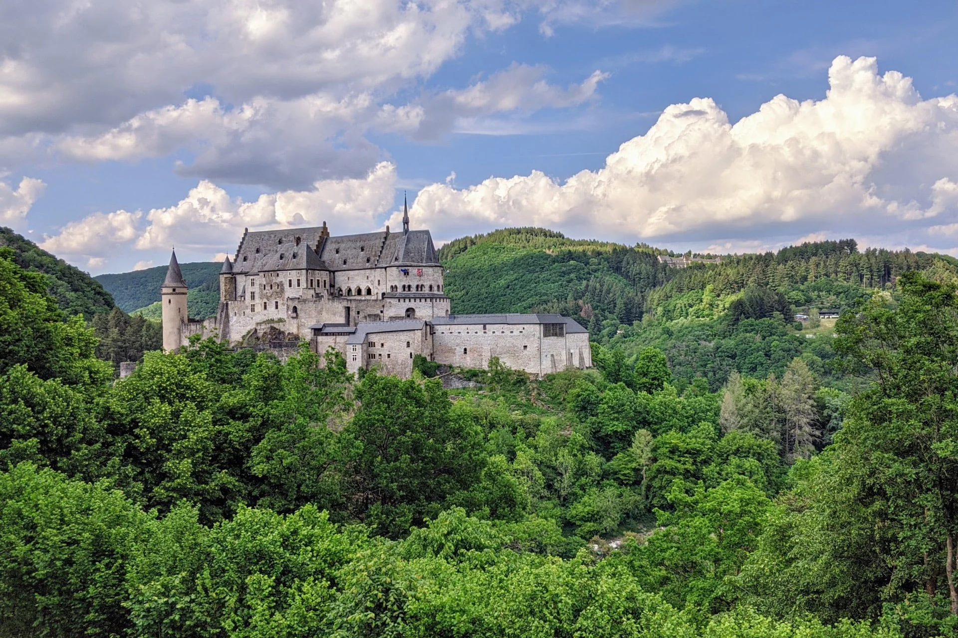 A castle on a hill surrounded by trees with vianden castle in the background.
