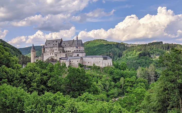 A castle on a hill surrounded by trees with vianden castle in the background.