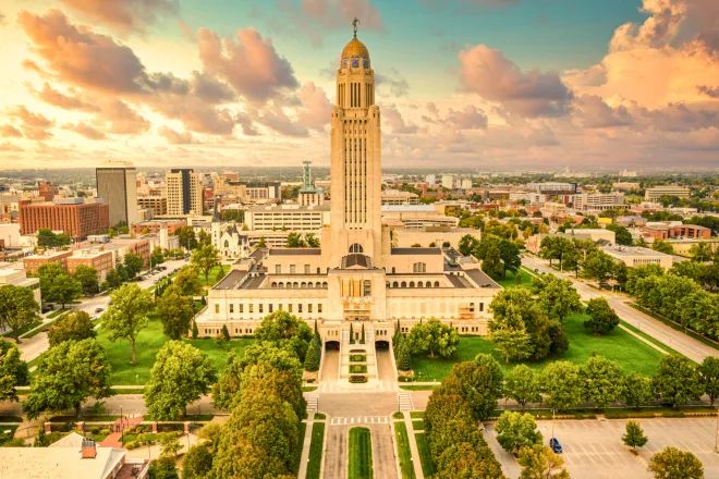 Aerial view of nebraska state capitol building in lincoln, surrounded by green landscape and urban cityscape under a cloudy sky.