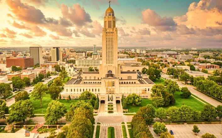 Aerial view of nebraska state capitol building in lincoln, surrounded by green landscape and urban cityscape under a cloudy sky.