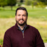 A man in a burgundy sweater smiles in front of a field.