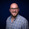 A person with a beard, smiling, wearing glasses and a checkered shirt, standing against a dark blue background.