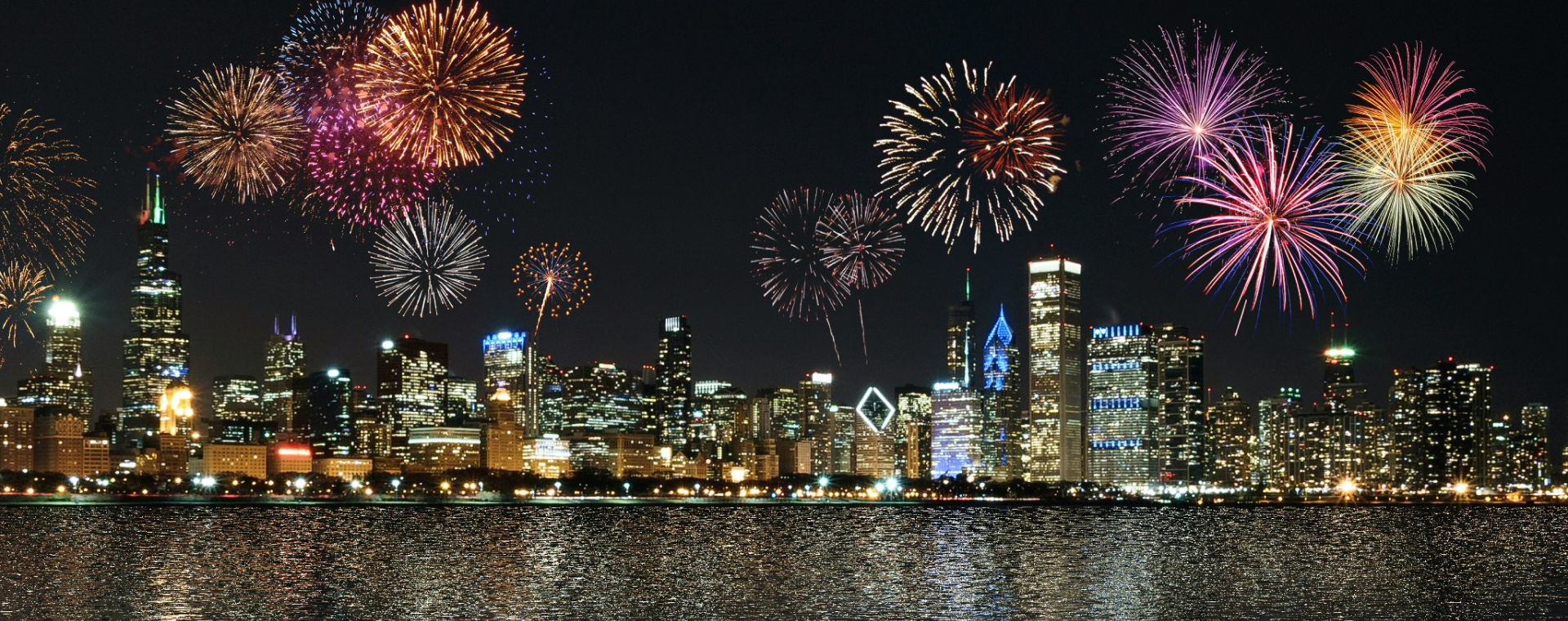 A colorful fireworks display over a city skyline at night, reflecting on the water below.