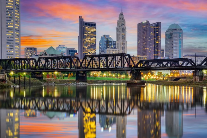 A vibrant sunset over a city skyline with modern high-rises and a historic truss bridge reflected in the calm river below.