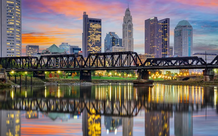 A vibrant sunset over a city skyline with modern high-rises and a historic truss bridge reflected in the calm river below.