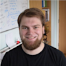 A man with a beard smiling in front of a desk.