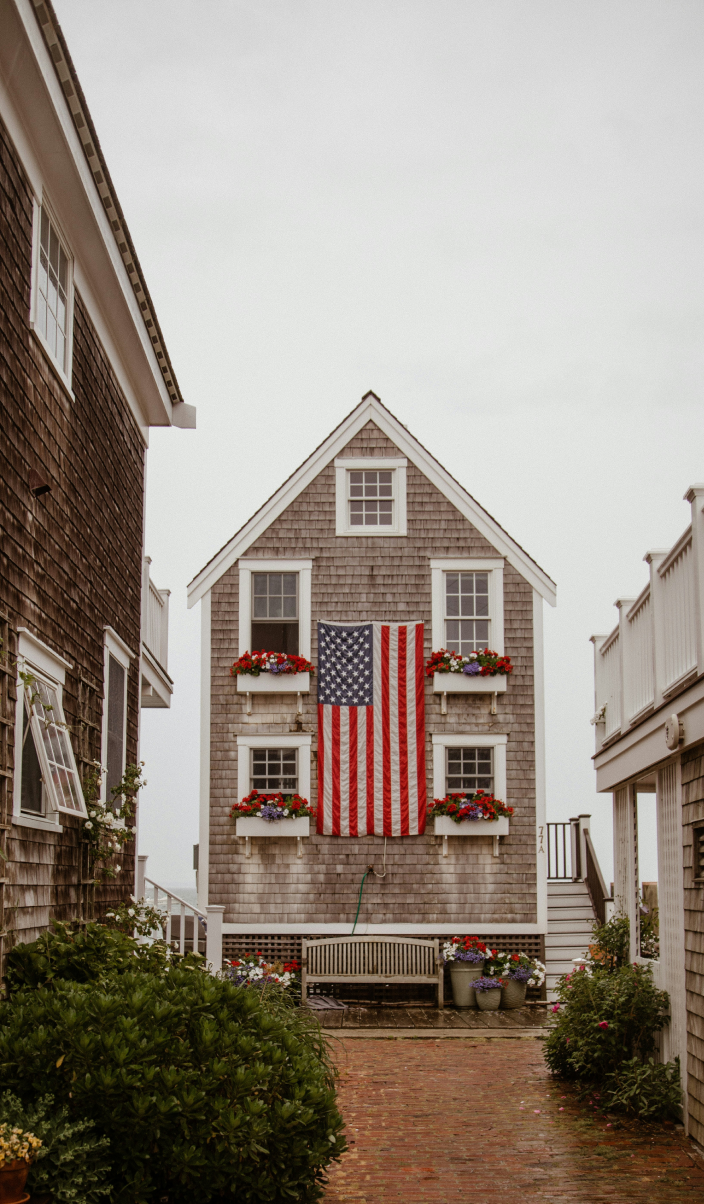 A traditional wooden house with an american flag hanging between two windows adorned with red flowers, on an overcast day.