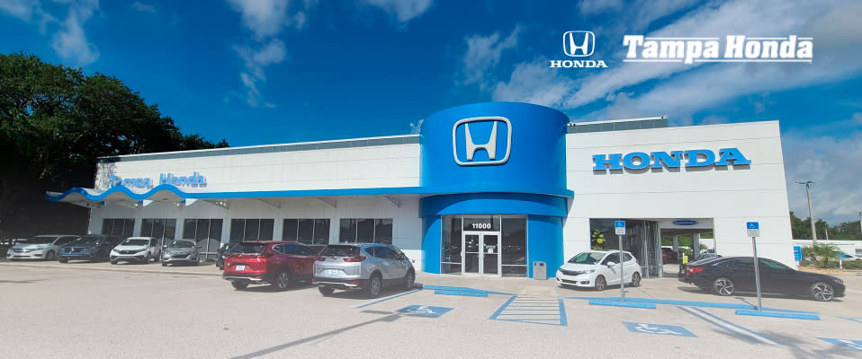 Photo of the Tampa Honda dealership storefront with a blue sky and parking lot containing a few cars. The building is grey with a large blue door including the Honda logo, and the company logo in in the right corner.