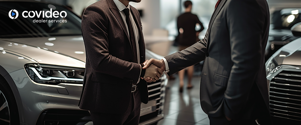 Two businessmen shaking hands in a car dealership, representing the trust and credibility built through customer testimonial videos. Covideo Dealer Services logo displayed in the top left corner, highlighting the service that facilitates the creation of impactful testimonial videos.