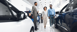 A photo of a man in a suit holding a clipboard in a dealership showroom, guesturing toward a car. A couple holding hands is looking at him and smiling.