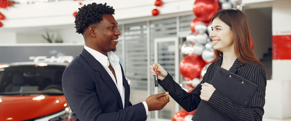 A woman holding a clipboard in a suit hands car keys to a man in a suit. They are both smiling and standing in a dealership showroom in front of a red car with balloons.