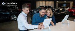 A car dealership staff member assists a couple using a tablet device inside the showroom. The couple looks engaged and happy as they explore options on the screen. The image prominently highlighting the use of advanced technology in the car-buying experience.