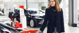 A photo of a woman in a blazer smiling has she looks at a new car in a dealership showroom