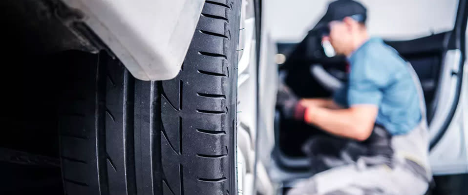A photo focused on the back tire of a car with a man in the background kneeling by an open door conducting a repair