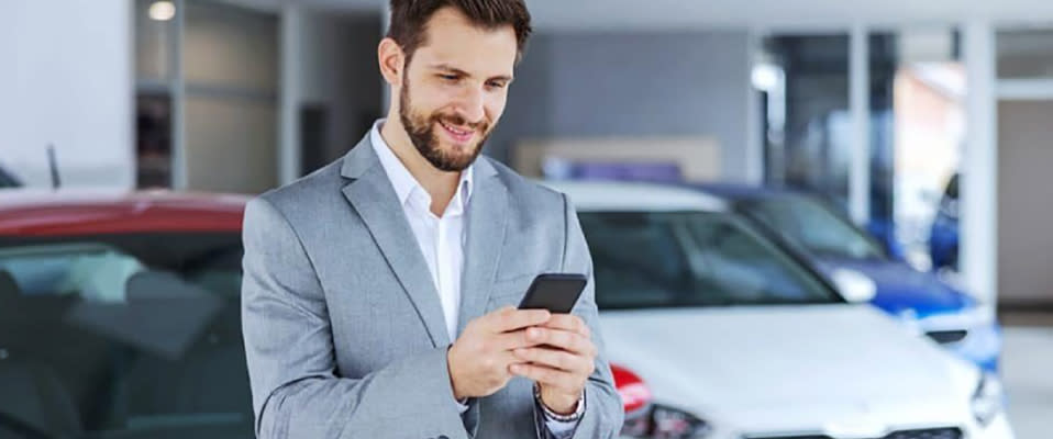 A photo of a man in a suit standing in front of a row of cars at a dealership, looking and smiling at his smartphone