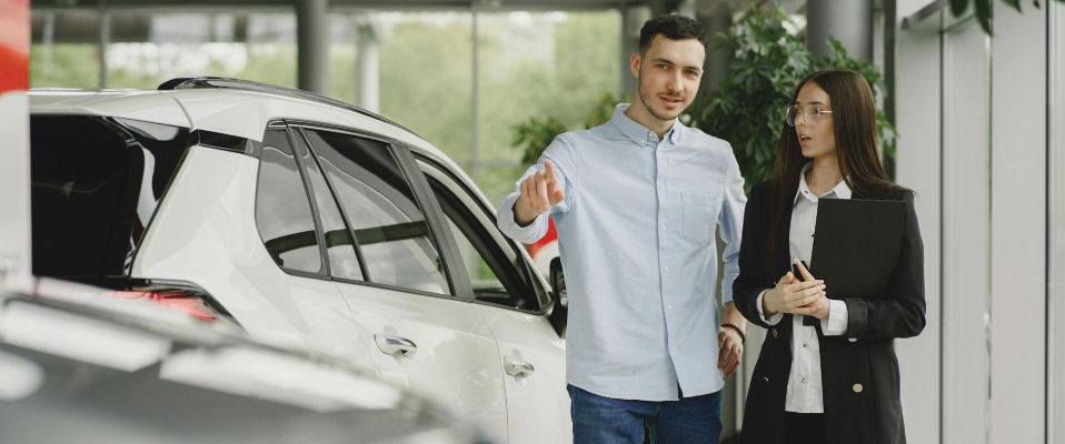 A photo of a man and a woman in a dealership. The man points at a car in the distance while the woman next to him listens and holds a folder.