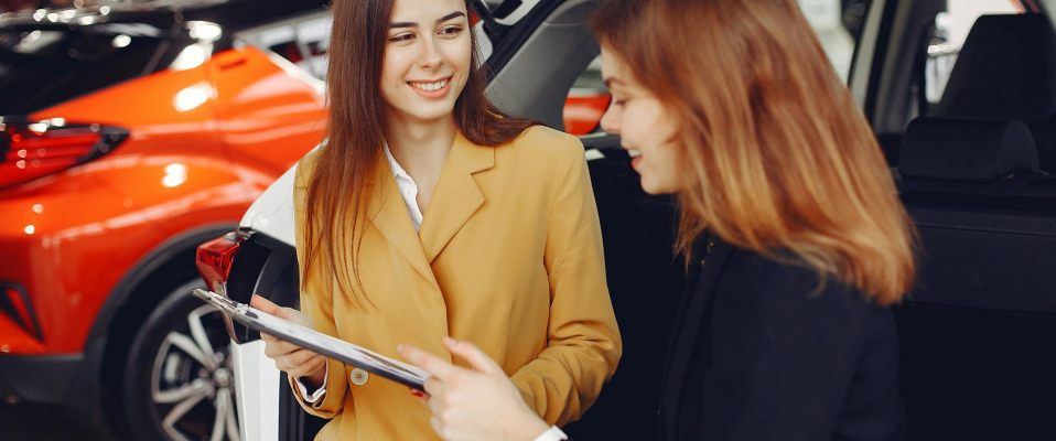 Two women in blazers smile and hold a clipboard together in front of a row of cars
