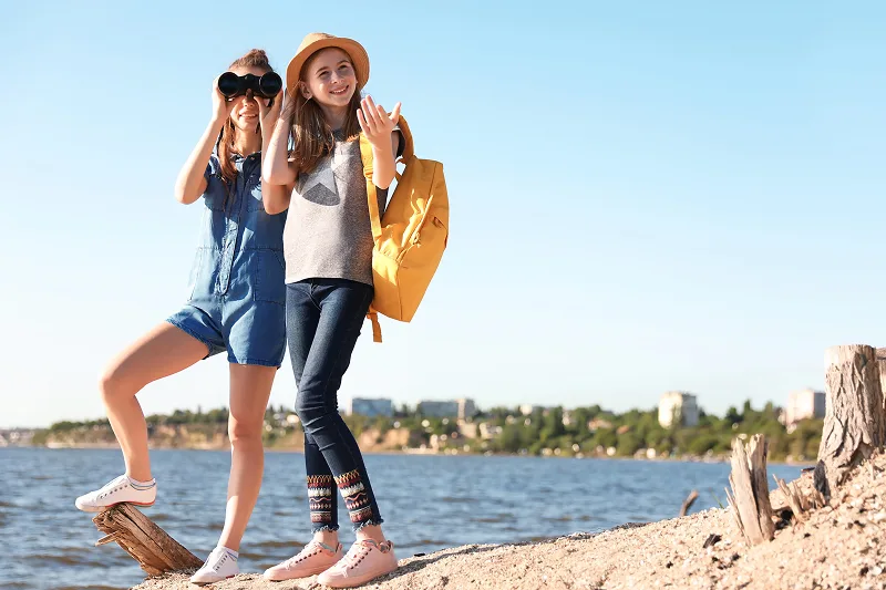 Dos chicas de pie a la orilla del mar, mientras una mira a través de unos binoculares.