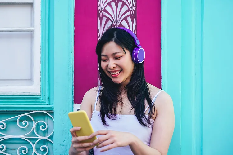 Chica con auriculares mirando su teléfono frente a una pared turquesa y morada.