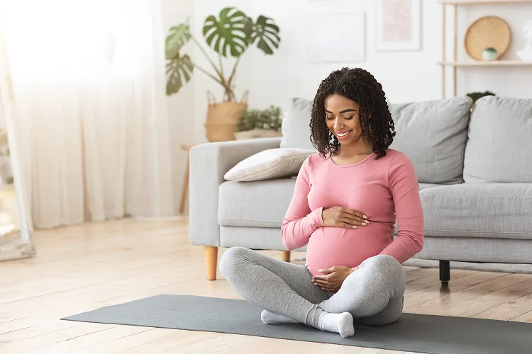 Mulher negra grávida sorridente abraçando a barriga, sentada em um tapete de exercícios.